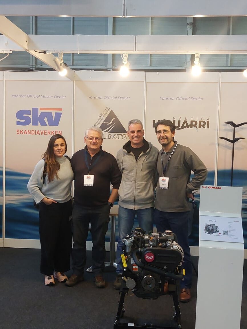 Four people wearing lanyards standing next to a display of an engine, likely at a trade show or exhibition.