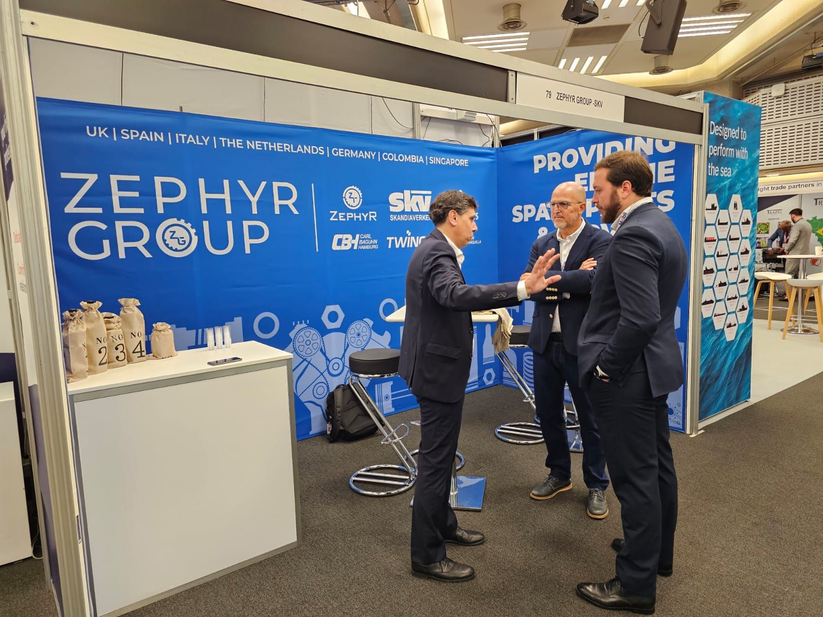 Three businessmen in suits engaged in conversation at a trade show booth with a blue and white Zephyr Group display background.