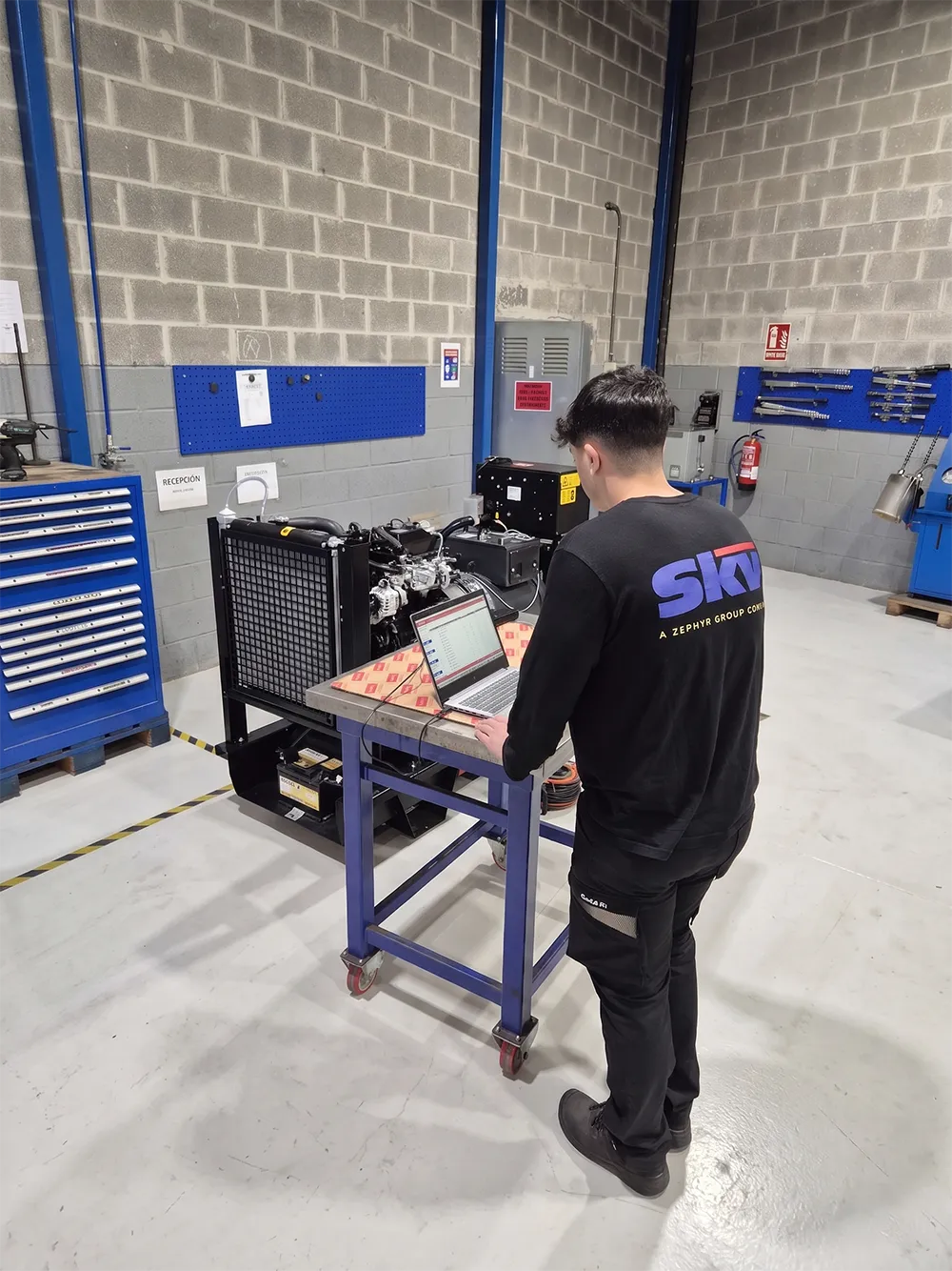 A mechanic in a black uniform inspecting equipment. The workshop has various tools and machinery.