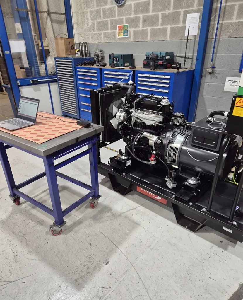 A laptop on a table next to a large industrial engine, with toolboxes in the background. The setup appears to be for engine testing or diagnostics.