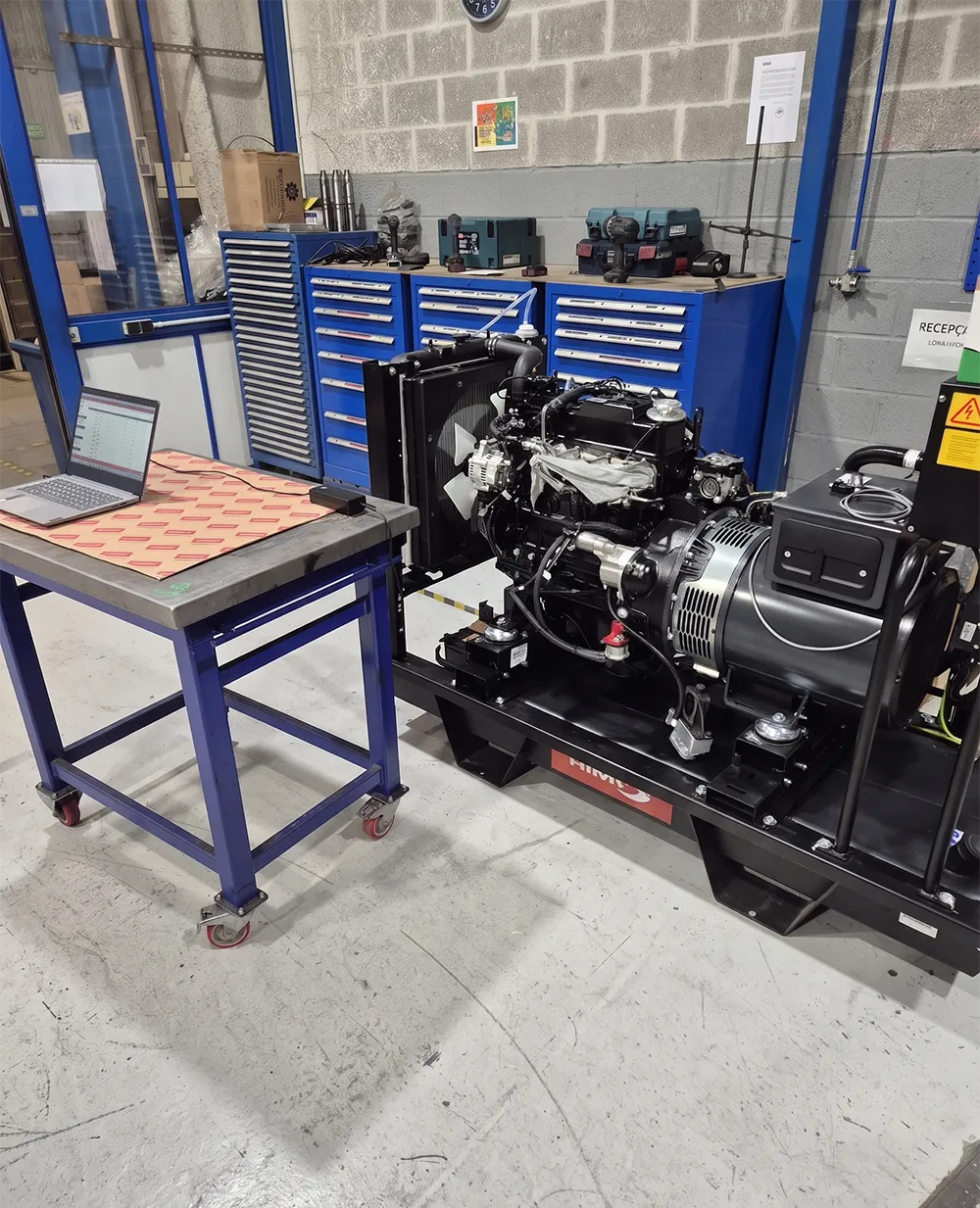 A laptop on a table next to a large industrial engine, with toolboxes in the background. The setup appears to be for engine testing or diagnostics.